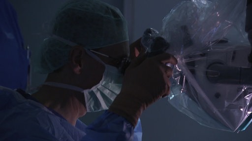 Ein Arzt der durch ein Mikroskop schaut. Er befindet sich in einem OP und trägt Op-Kleidung.