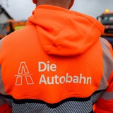 Ein Mensch in oranger Jacke steht vor einem Baustellenfahrzeug.