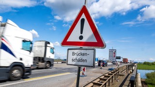Auf der Weserbrücke der A1 in Bremen fahren Lastwagen. Im Vordergrund ist ein Warnschild mit dem Hinweis "Brückenschäden" zu sehen.