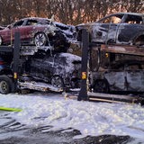 Ein ausgebrannter Autotransporter steht auf einer Autobahn. Weißer Löschschaum liegt auf dne Autos und auf der Fahrbahn.