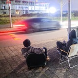 Ein Auto rast durch eine Straße, beobachtet von drei jungen Männern