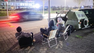Ein Auto rast durch eine Straße, beobachtet von drei jungen Männern