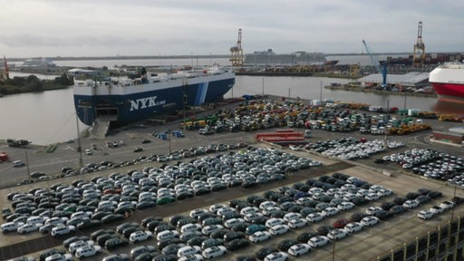 Hunderte Autos warten in einem Hafen darauf auf ein großes Containerschiff verladen zu werden