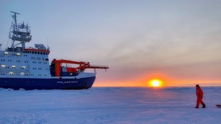 28.8.: Die Polarstern bei tiefstehender Sonne im Eis, vorne ein Besatzungsmitglied mit einem Schlitten