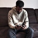 Der Mann Ayyub sitzt auf einem Sofa und schaut auf ein Handy.