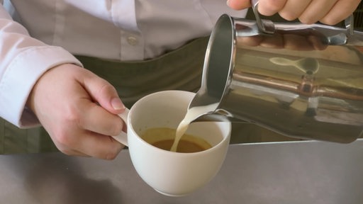 Eine Person bereitet Kaffee zu und gießt Milch in die weiße Tasse.