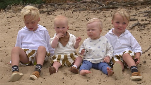 Vier Kleinkinder sitzen nebeneinander im Sand an einem Strand.