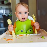 Ein Baby sitzt auf einem Hochstuhl und hält einen Löffel und eine Gabel in der Hand.