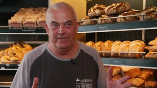 Eine Person steht in einem Laden, es sieht aus wie eine Bäckerei. Hinter ihm sind Brote und Brötchen zu erkennen.