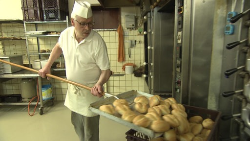 Ein Bäcker holt gerade frische Brötchen auf einer großen Schaufel aus dem Ofen.
