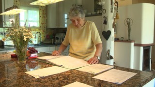 Eine ältere Frau steht am Tisch in ihrer Küche und schaut auf Dokumente.