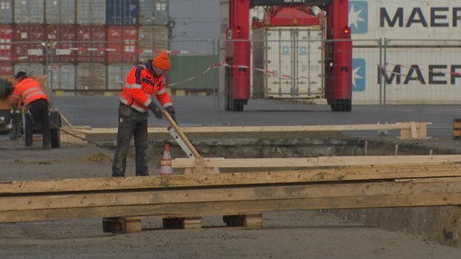 Zwei Bauarbeiter stehen auf einer Baustelle vor gestapelten Containern.