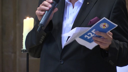 Ein Mann hält ein Papier mit der Aufschrift "125 Bahnhofsmission" und ein Mikrofon in der Hand