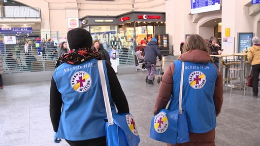 Zwei Frauen im Bahnhof mit Kleidung und Logos der Bahnhofsmission. 
