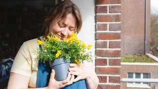 Eine Frau mit Schürze auf einem Balkon hält einen Topf mit Blumen hoch und riecht daran und lächelt (Symbolbild)