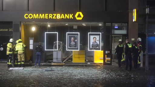 Eine zersprängte Glasscheibe und kaputte Geldautomaten von einer Bankfiliale, Polizisten stehen davor