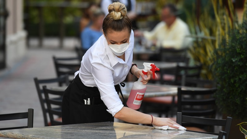  Eine Kellnerin mit Mund-Nasenschutz wischt Tische einer Außengastronomie sauber