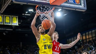 Der Basketball-Profi Norris Agbakoko von den Baskets Oldenburg hängt am Korb, nachdem er den Ball darin versenkt hat, sein Gegenspieler Justus Hollatz von Bayern München springt vergeblich hoch.
