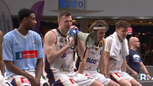 Basketballer der Eisbären Bremerhaven um Robert Oehle sitzen nebeneinander nach dem Sieg mit einem Megaphon und singen mit den Fans.