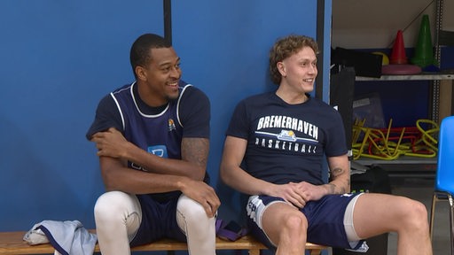 Zwei Spieler der Basketballmannschaft Eisbären Bremerhaven sitzen nebeneinander in einer Sporthalle auf einer Bank.