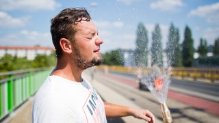 ein Mann steht auf einer Baustelle und sprüht sich mit einem Schlauch Wasser ins Gesicht