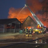 Feuerwehr-Leute stehen vor einem brennenden Bauernhof, die Flammen lodern.