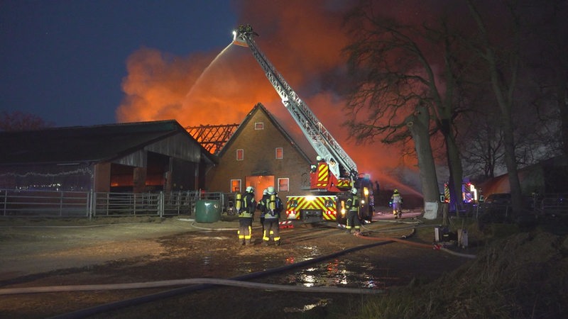 Feuerwehr-Leute stehen vor einem brennenden Bauernhof, die Flammen lodern.