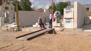 Zwei Personen unterhalten sich auf einer Baustelle in einem Rohbau eines Hauses. 