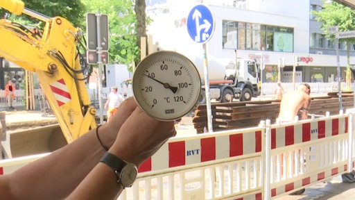 Eine Person hält ein Thermometer neben einer Baustelle hoch. Die Anzeige liegt fast bei 40 Grad Celsius.