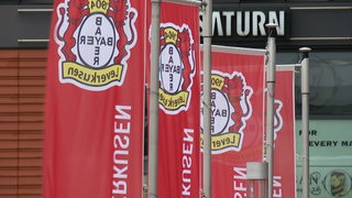 Es sind drei Flaggen von Bayer Leverkusen vor einem Gebäude zu sehen.