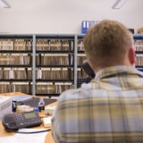 Ein Beamter sitzt am Schreibtisch und telefoniert, im Hintergrund Regale mit Akten (Symbolbild)