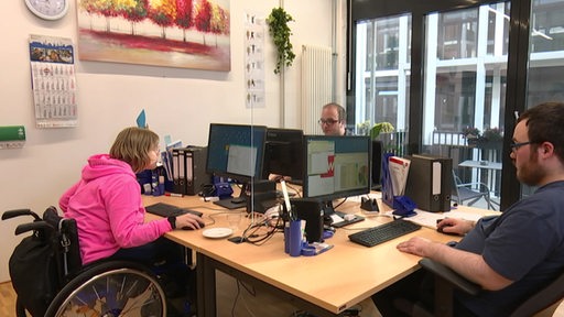 Ein Büro, in dem zwei Männer und eine Frau im Rollstuhl arbeiten.