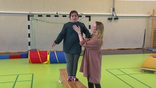 Ein dunkelhaariges Mädchen balanciert auf einer Bank, eine blonde Frau hilft ihr dabei. 