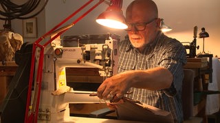 Bernhard Kiesling näht Leder an seiner Industrienähmaschine zusammen. Eine rote Lampe spendet ihm dabei Licht. 