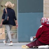 Eine Frau mit Kopftuch bettelt mit einem Pappbecher um Geld. Im Hintergrund eine Frau mit Shopping-Tüte.