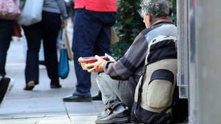 Ein mittelalter Mann sitzt auf dem Gehweg und hält Passanten ein Körbchen entgegen.