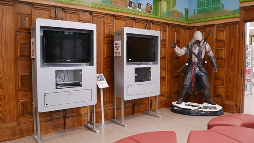 Zwei Fernseher mit Playstations stehen neben einer lebensgroßen Actionfigur in einem Raum der Bibliothek.