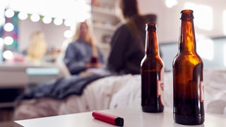 Zwei offene Bierflaschen stehen auf einem Tisch, im Hintergrund unterhalten sich zwei junge Frauen