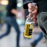 Ein Jugendlicher steht mit einer Flasche Bier in der Hand in einem Bahnhof, im Hintergrund gehen Menschen.