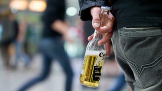 Ein Jugendlicher steht mit einer Flasche Bier in der Hand in einem Bahnhof, im Hintergrund gehen Menschen.