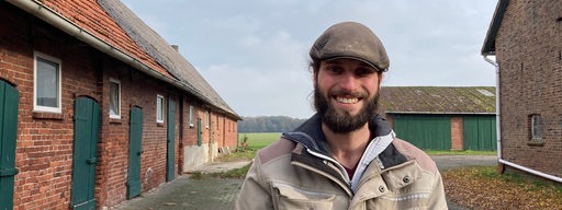 Ein Mann mit Bart und Mütze steht auf einem Bauernhof.