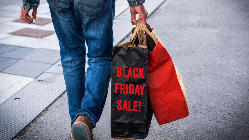 Ein Mann geht mit Einkaufstüten die Straße entlang. Auf einer schwarzen Tüte steht: Black Friday Sale.