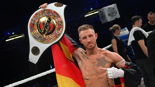 Boxer Pascal Schroth, der in eine Deutschland-Fahne gehüllt ist, hält seinen Weltmeister-Gürtel in dei Höhe.