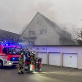 Zwei Feuerwehrleute stehen vor einem Einsatzfahrzeug. Im Hintergrund ist ein brennendes Haus zu sehen.