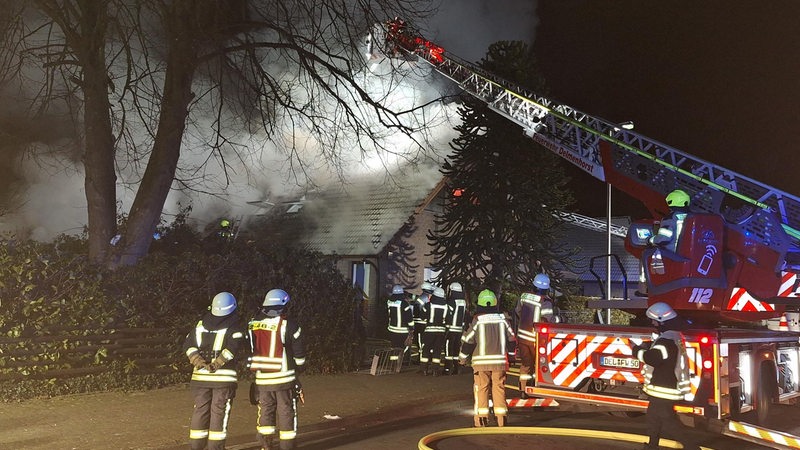 mehrere Feuerwehrleute stehen vor einem Wohnhaus mit Rauchschwaden, von der ausgefahrenen Drehleiter eines Einsatzwagens der Berufsfeuerwehr Delmenhorst aus wird Löschwasser versprüht