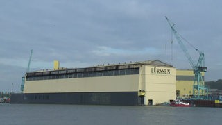 Die gelbliche Halle der Lürssen Werft und ein grünlicher Kran sind zu sehen.