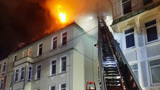 Ein Feuerwehrauto mit ausgefahrener Drehleiter steht vor einem Mehrfamilienhaus. Das Dach brennt.