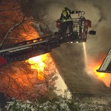 Flammen schlagen aus den Fenstern eines Hauses, ein Feuerwehrmann steht auf der Drehleiter und löscht es.
