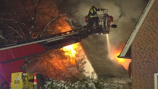 Flammen schlagen aus den Fenstern eines Hauses, ein Feuerwehrmann steht auf der Drehleiter und löscht es.