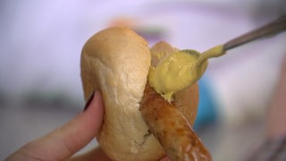 Eine Hand hält ein Brötchen mit einer Bratwurst drinnen und schmiert mit einem Löffel Senf drauf. 